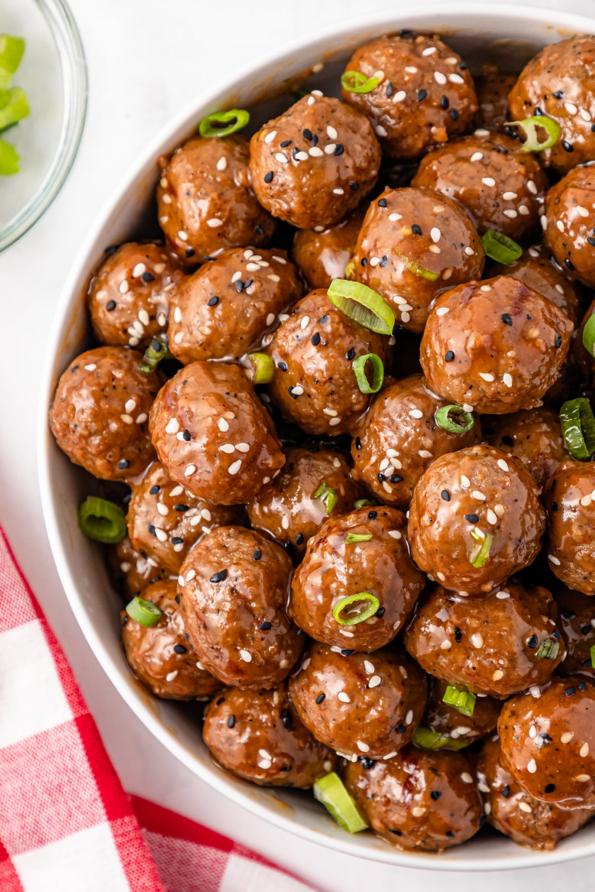 Bowl of glazed meatballs garnished with sesame seeds and chopped green onions, placed on a white surface with a red and white checkered cloth nearby.