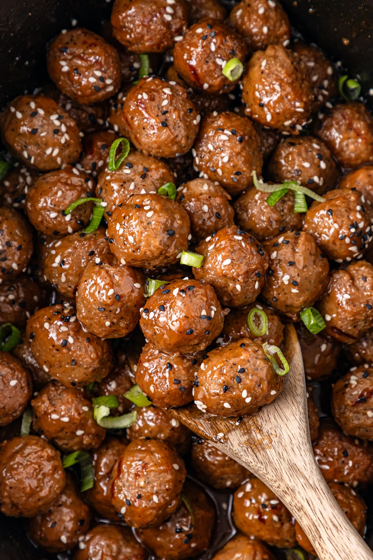 A wooden spoon scoops glazed meatballs garnished with sesame seeds and sliced green onions from a pan filled with more meatballs.