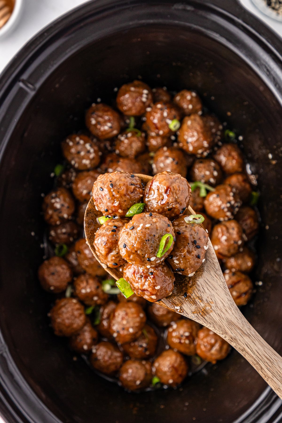 A wooden spoon holds several glazed meatballs topped with sliced green onions over a slow cooker filled with more meatballs.