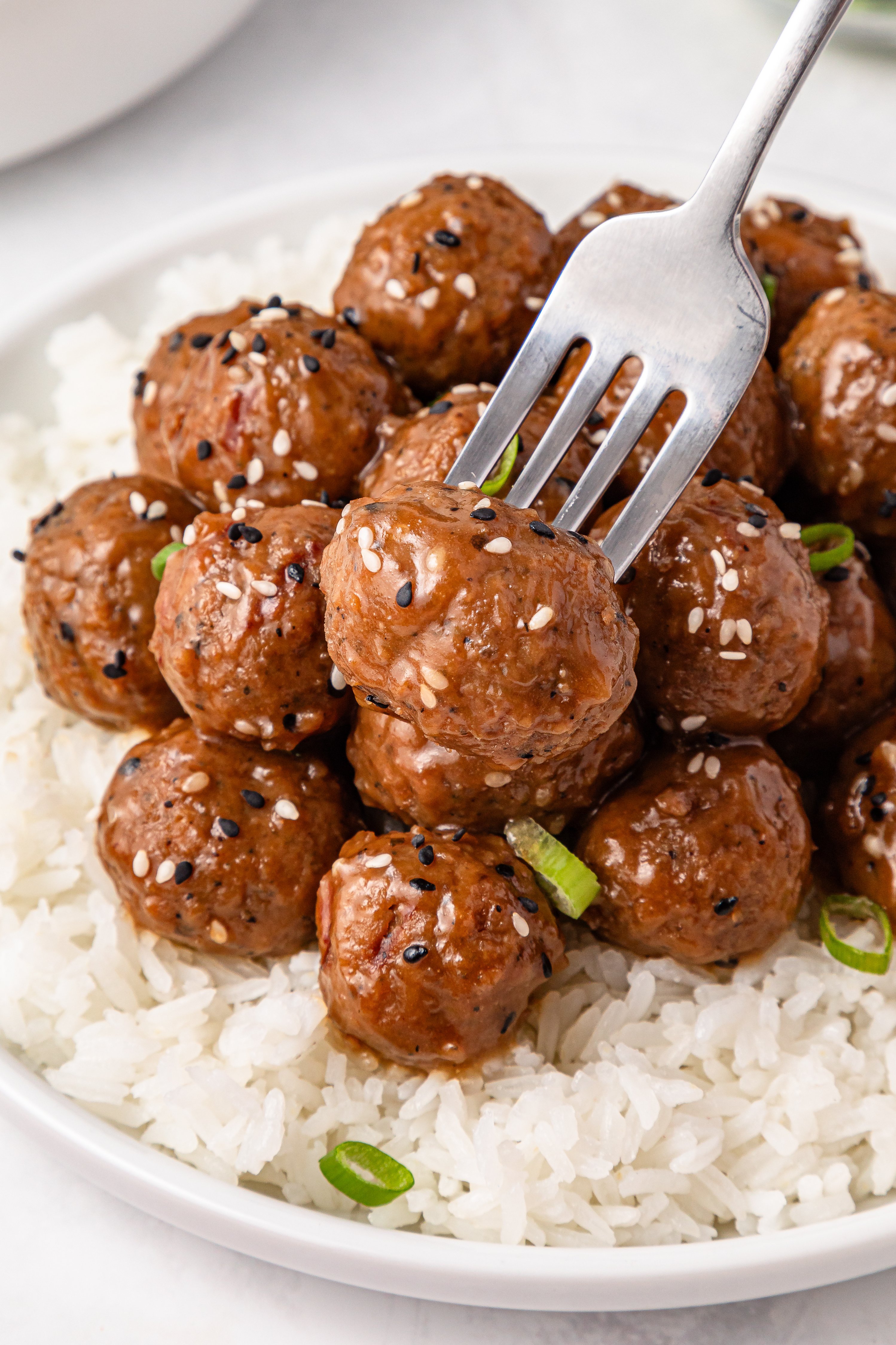 A plate of white rice topped with glazed meatballs sprinkled with sesame seeds and sliced green onions, with a fork picking up one meatball.