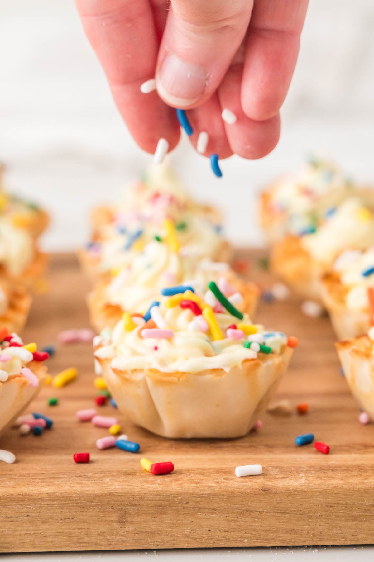 A hand sprinkles colorful sprinkles onto a mini pastry with cheesecake filling, with more decorated cheesecake cups on a wooden board in the background.