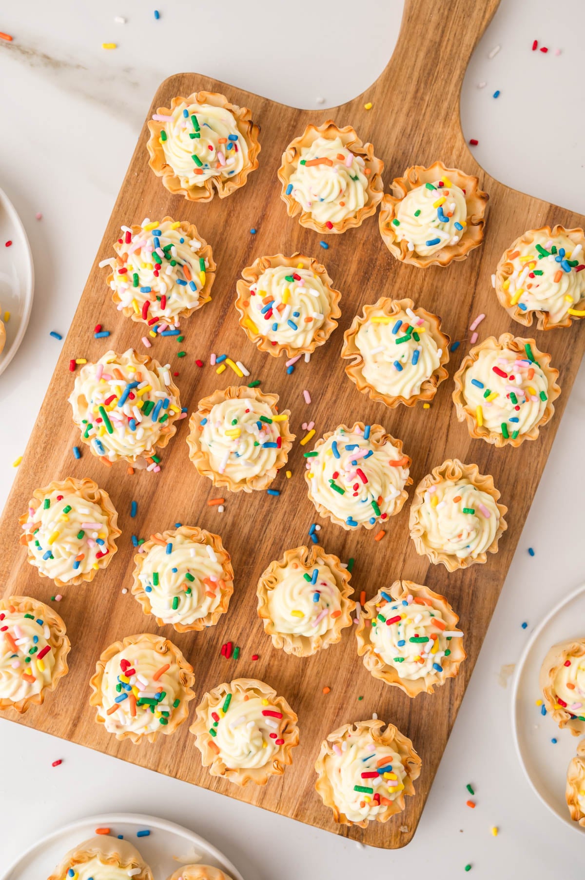 Mini tart shells filled with cheesecake and topped with colorful sprinkles are arranged on a wooden serving board and some white plates.