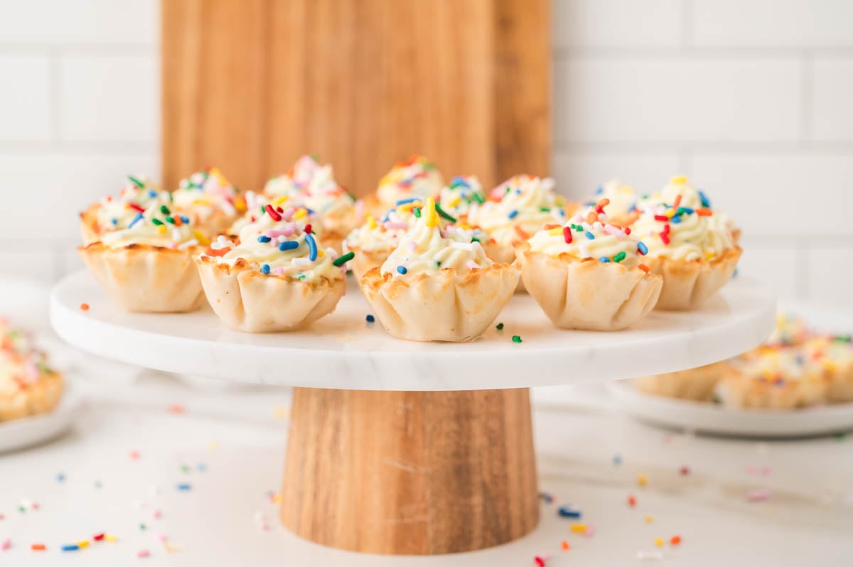 Mini phyllo shells filled with cheesecake and topped with rainbow sprinkles are displayed on a white cake stand with a wooden base.