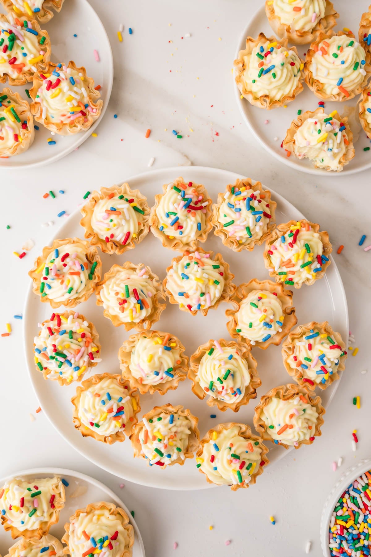 Mini tart shells filled with piped cheesecake and topped with colorful sprinkles are arranged on white plates and a white surface.