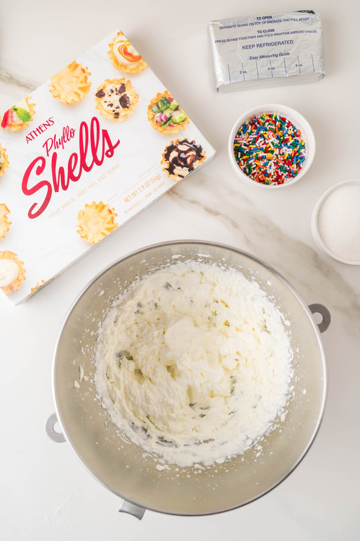 Metal bowl with whipped cream mixture, a box of phyllo shells, a bowl of sprinkles, a brick of cream cheese, and a bowl of sugar on a white countertop.