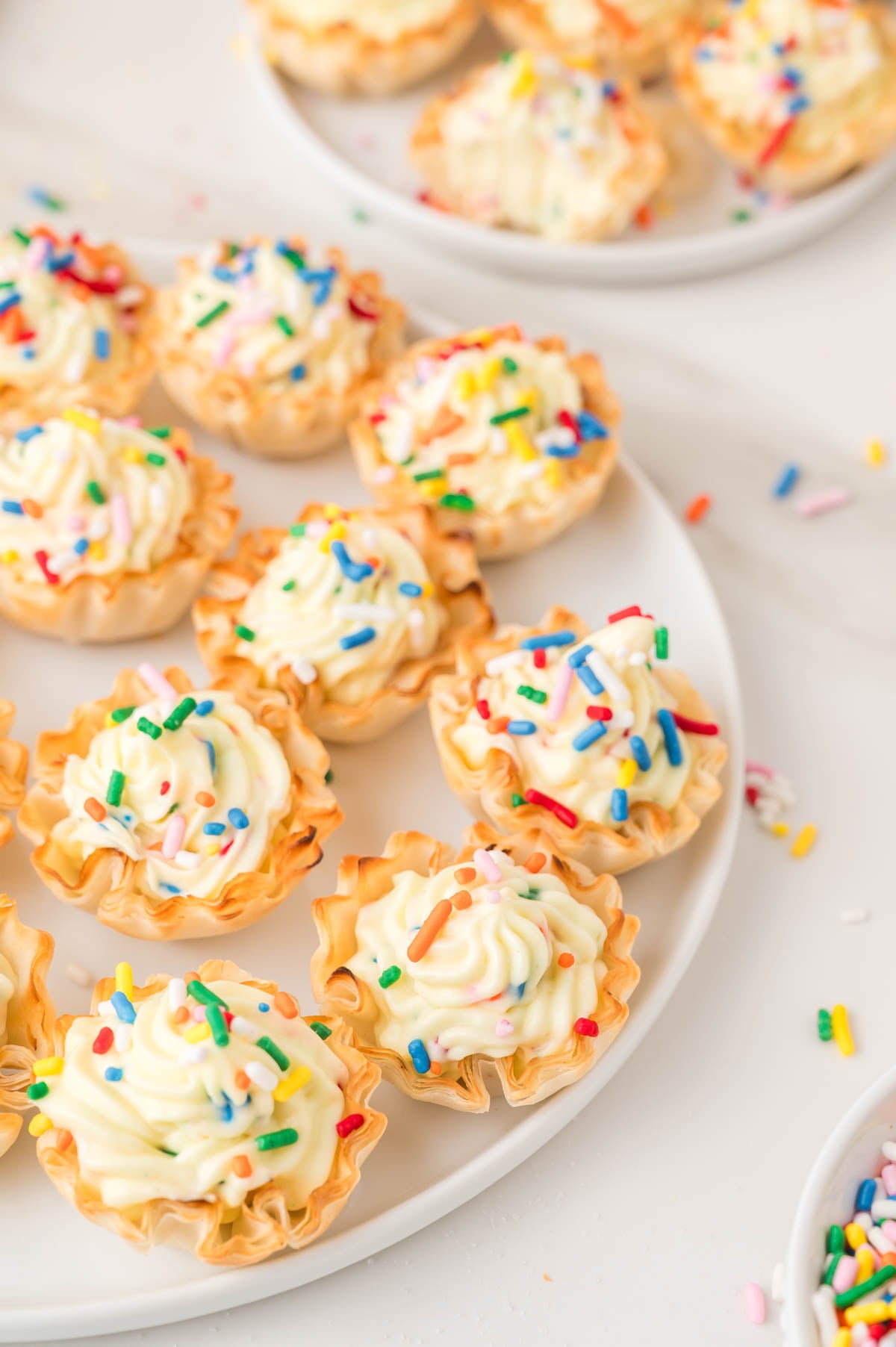 Mini pastry cups filled with cheesecake and topped with colorful sprinkles arranged on a white plate.