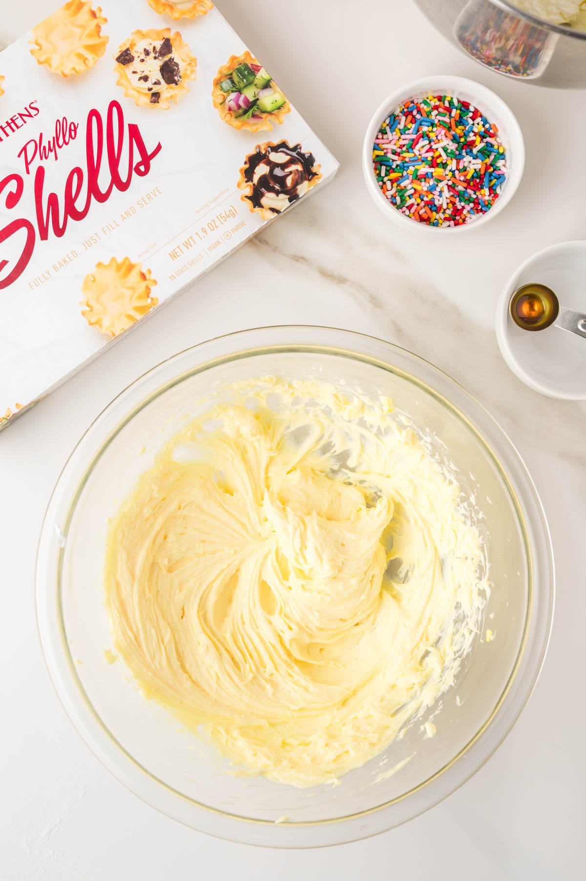 Bowl of whipped yellow batter, measuring spoon with vanilla, bowl of rainbow sprinkles, and a box of mini phyllo shells on a white surface.
