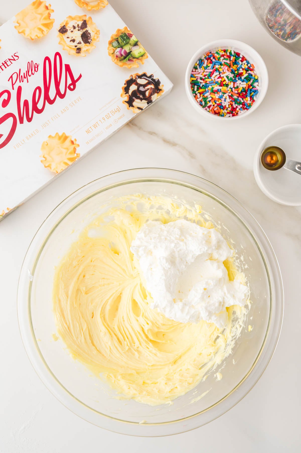 A glass bowl with whipped cream mixture, a box of phyllo shells, a dish of colorful sprinkles, and a measuring spoon with vanilla on a white countertop.