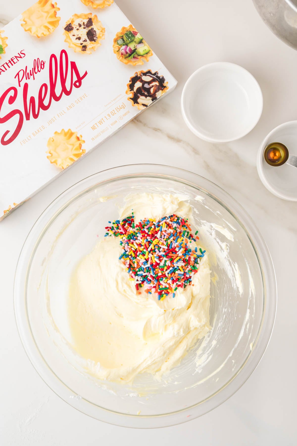A glass bowl of creamy white cheesecake mixture topped with colorful sprinkles sits on a white counter next to phyllo shells packaging and small bowls.