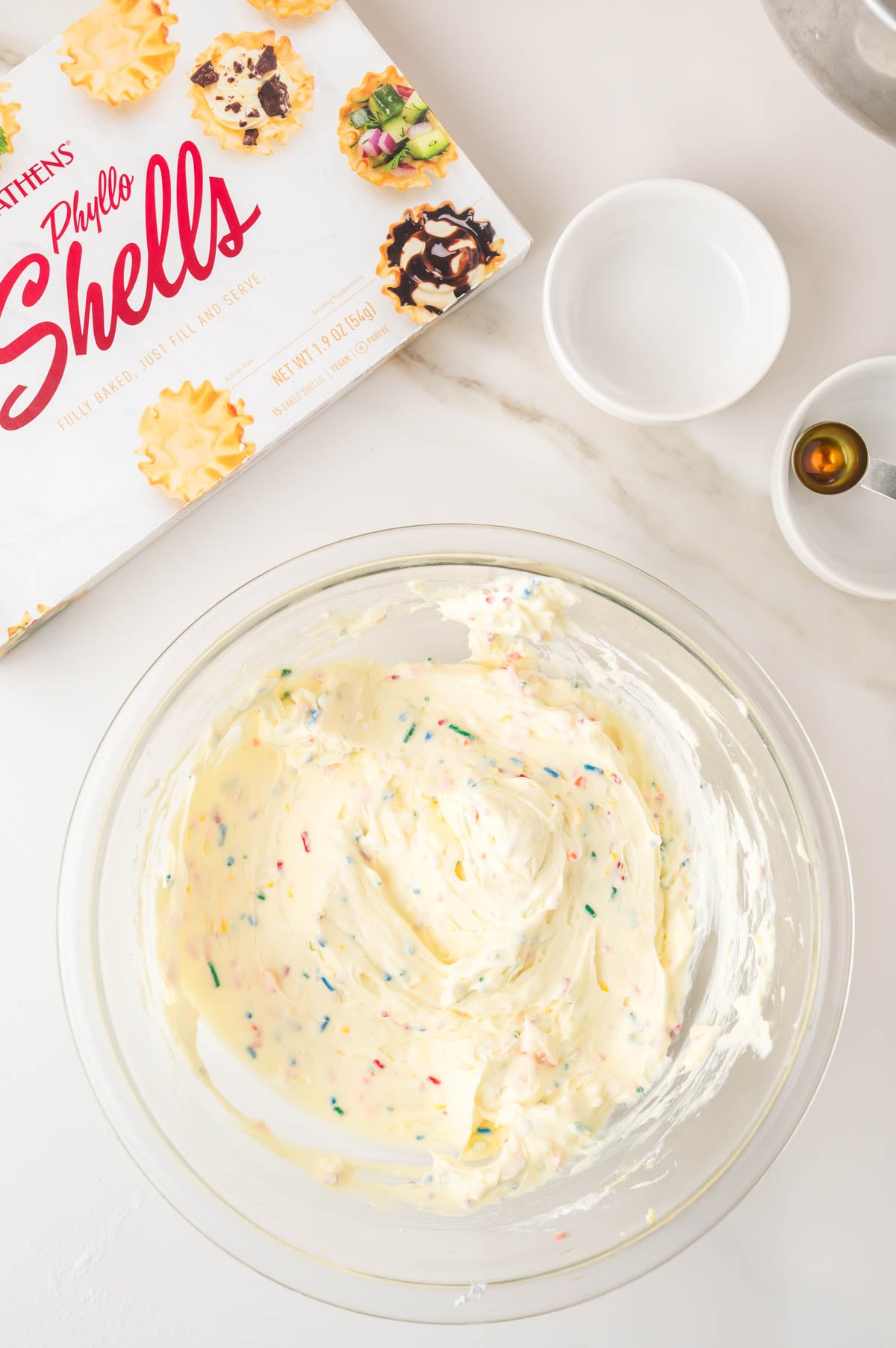 A glass bowl with sprinkled cream cheese mixture, a box of phyllo shells, a small bowl with vanilla extract, and an empty white dish on a marble surface.