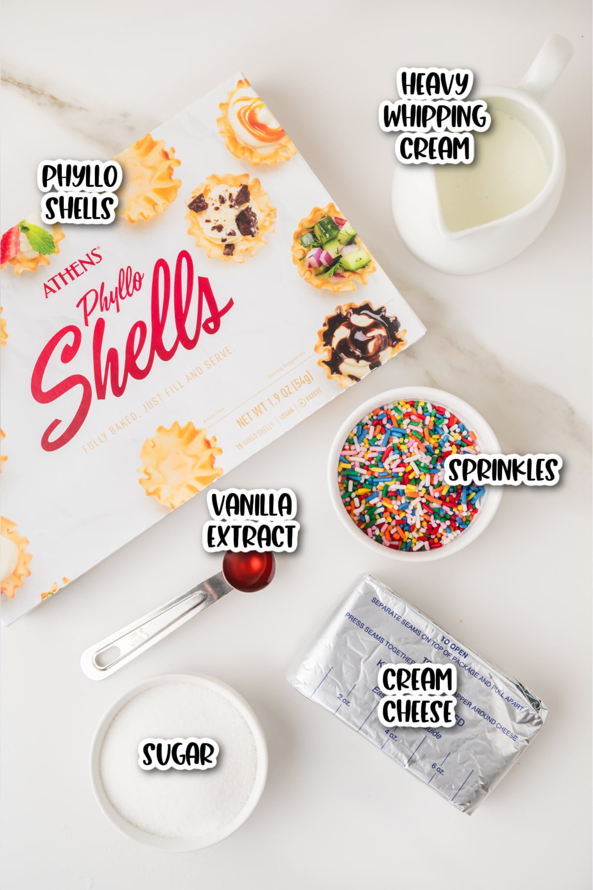 Box of phyllo shells, a cup of heavy whipping cream, a bowl of sprinkles, a package of cream cheese, a bowl of sugar, and a spoon of vanilla extract arranged on a white surface.