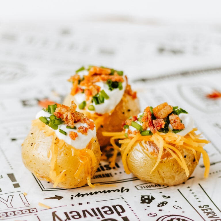 Three baked potatoes topped with sour cream, shredded cheese, chopped chives, and bacon bits are arranged on printed paper.