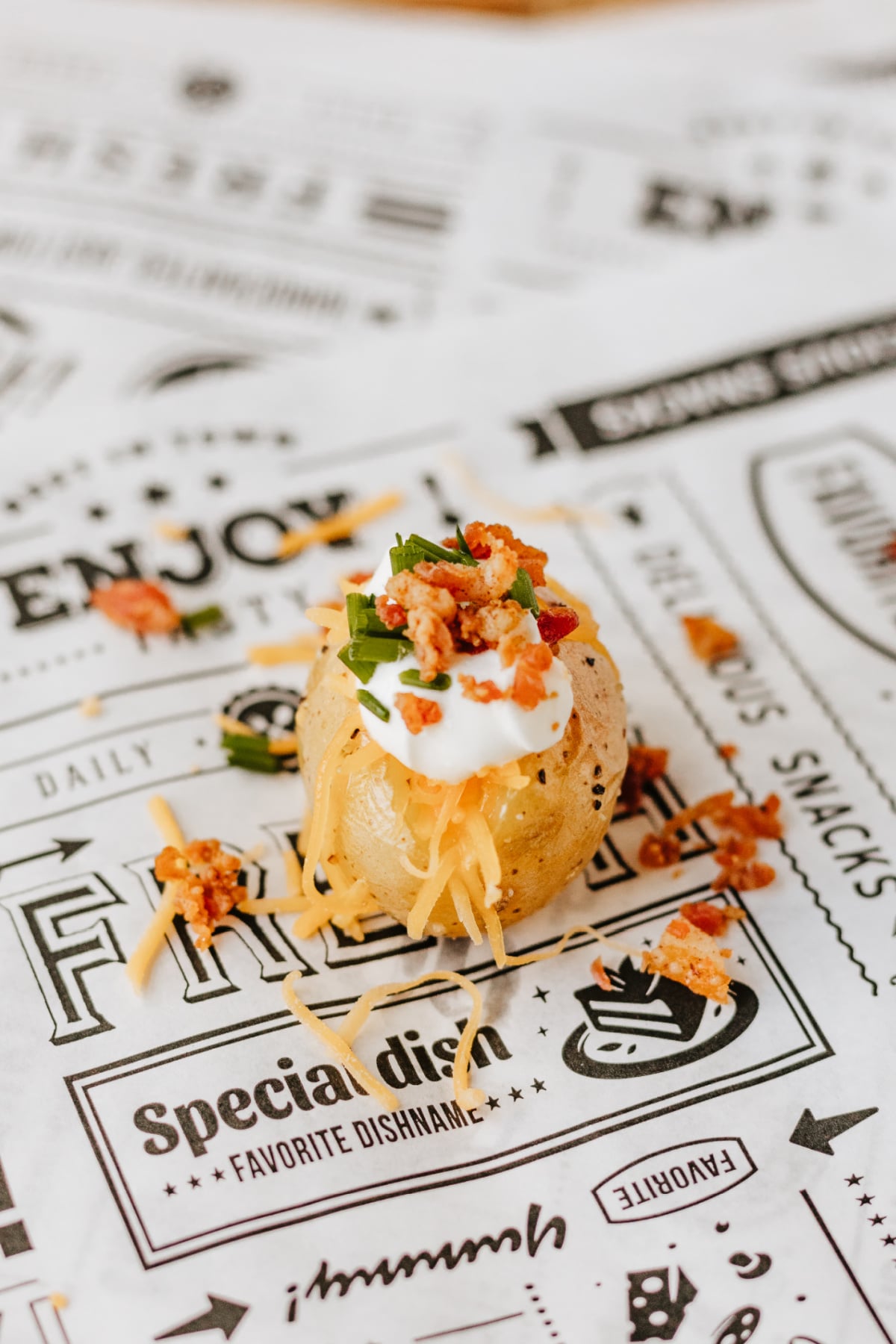 A small baked potato topped with sour cream, shredded cheese, bacon bits, and chopped herbs sits on decorative printed paper.