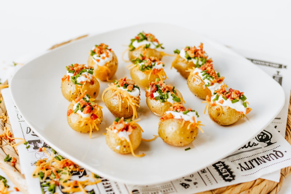 A white plate with small baked potatoes topped with sour cream, shredded cheese, chives, and bacon bits, arranged neatly on a table.