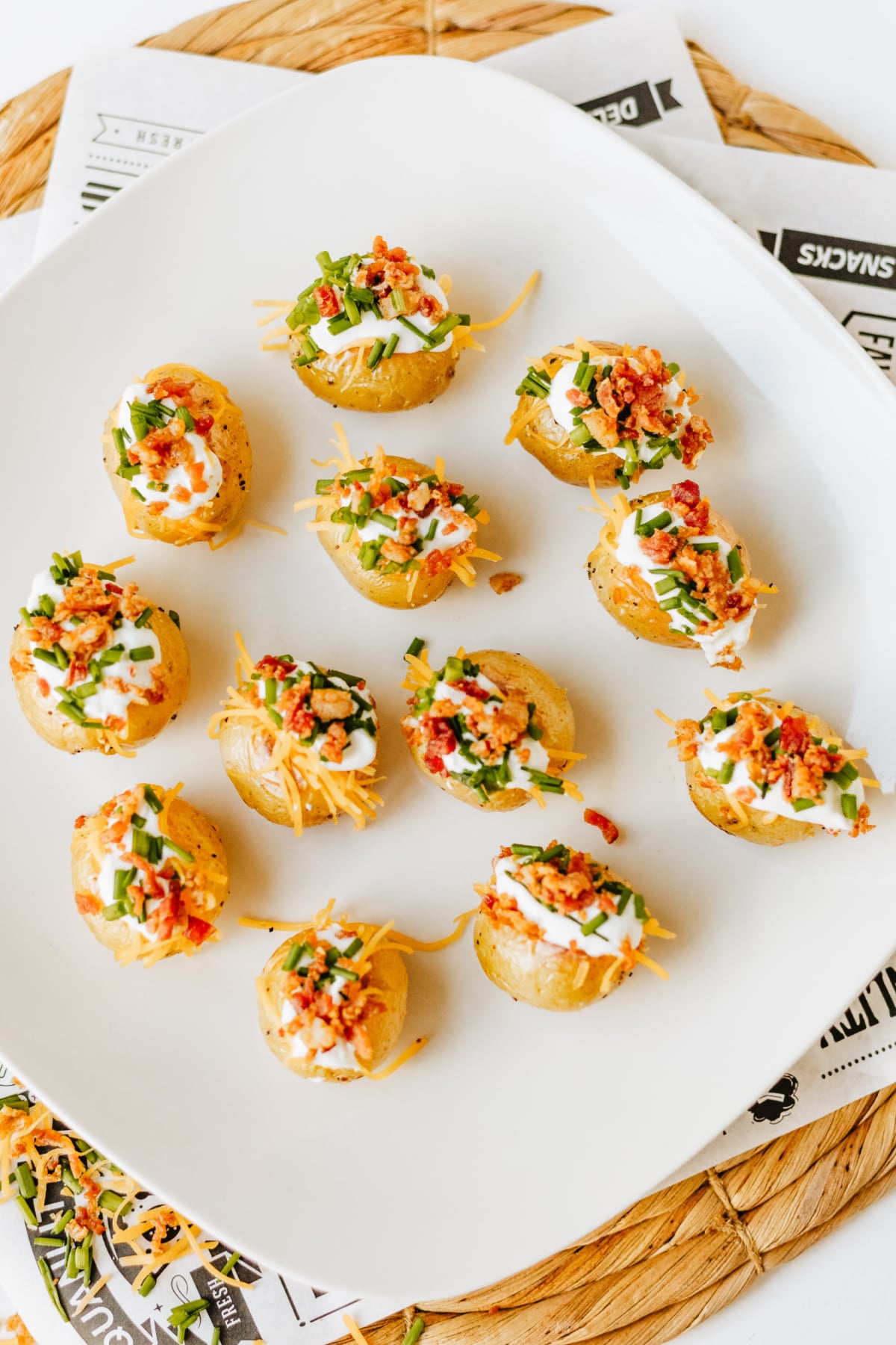 Small baked potatoes topped with sour cream, shredded cheese, chopped green onions, and bacon bits, arranged on a white square plate.