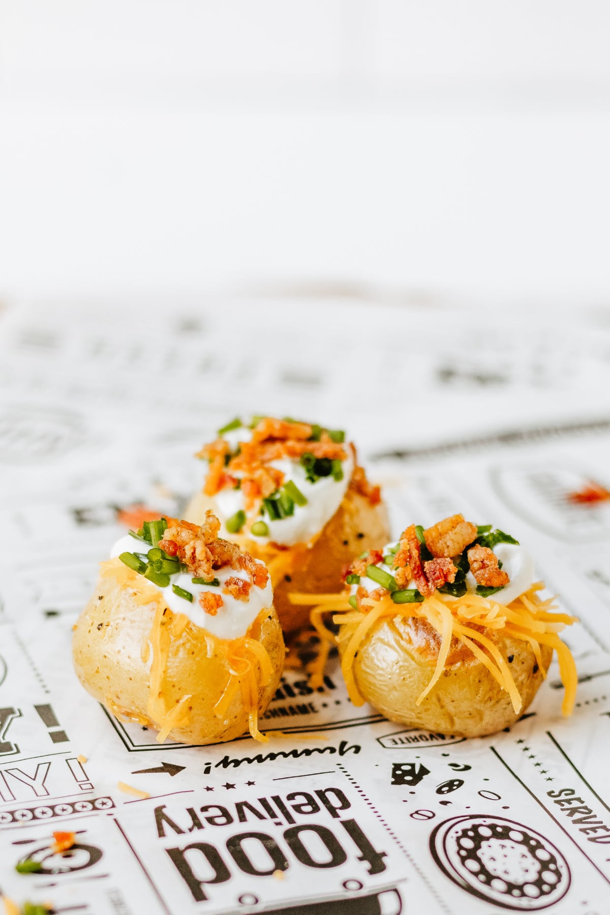 Three baked potatoes topped with sour cream, shredded cheddar cheese, bacon bits, and chopped chives, placed on paper with various food-themed graphics and text.