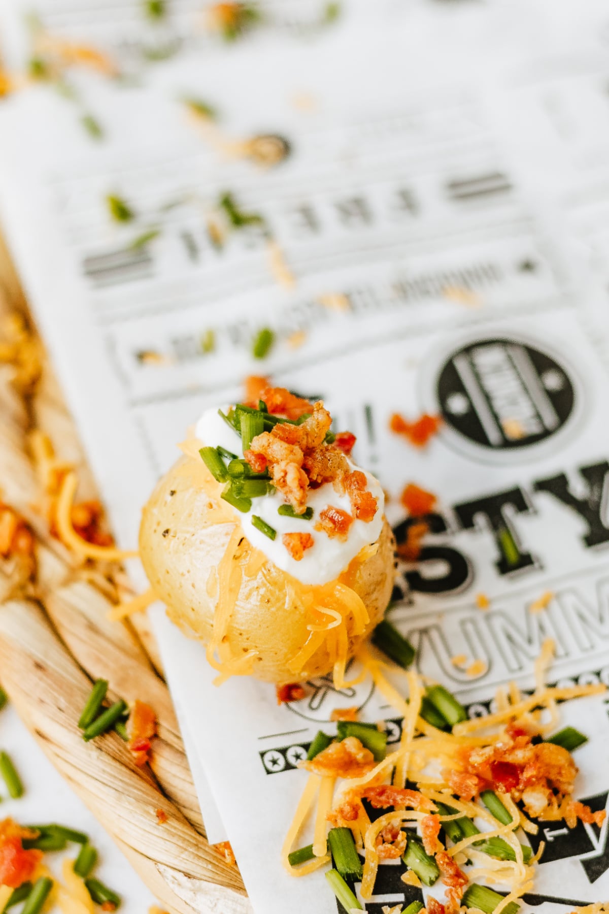 A baked potato topped with sour cream, shredded cheese, chopped chives, and bacon bits sits on decorative paper with scattered toppings around it.