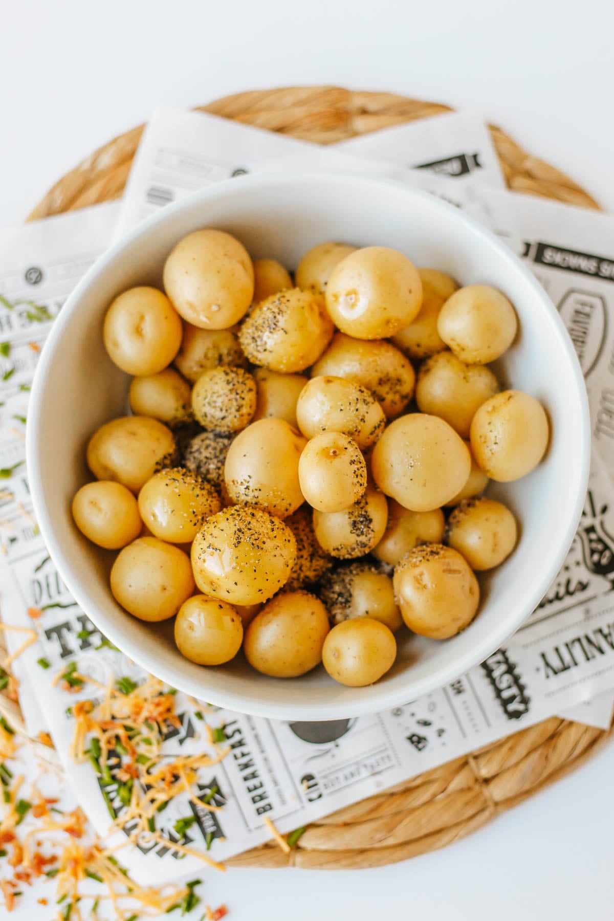 A white bowl filled with small potatoes topped with cracked black pepper, placed on a woven mat and printed paper.