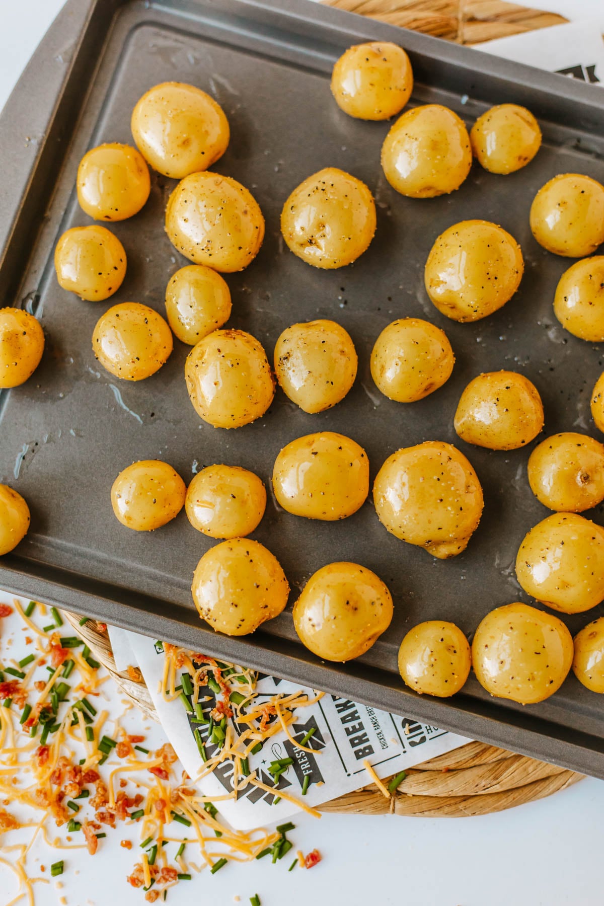 A baking tray with whole, seasoned baby potatoes, ready for roasting. Chopped herbs and shredded cheese are scattered on the surface nearby.