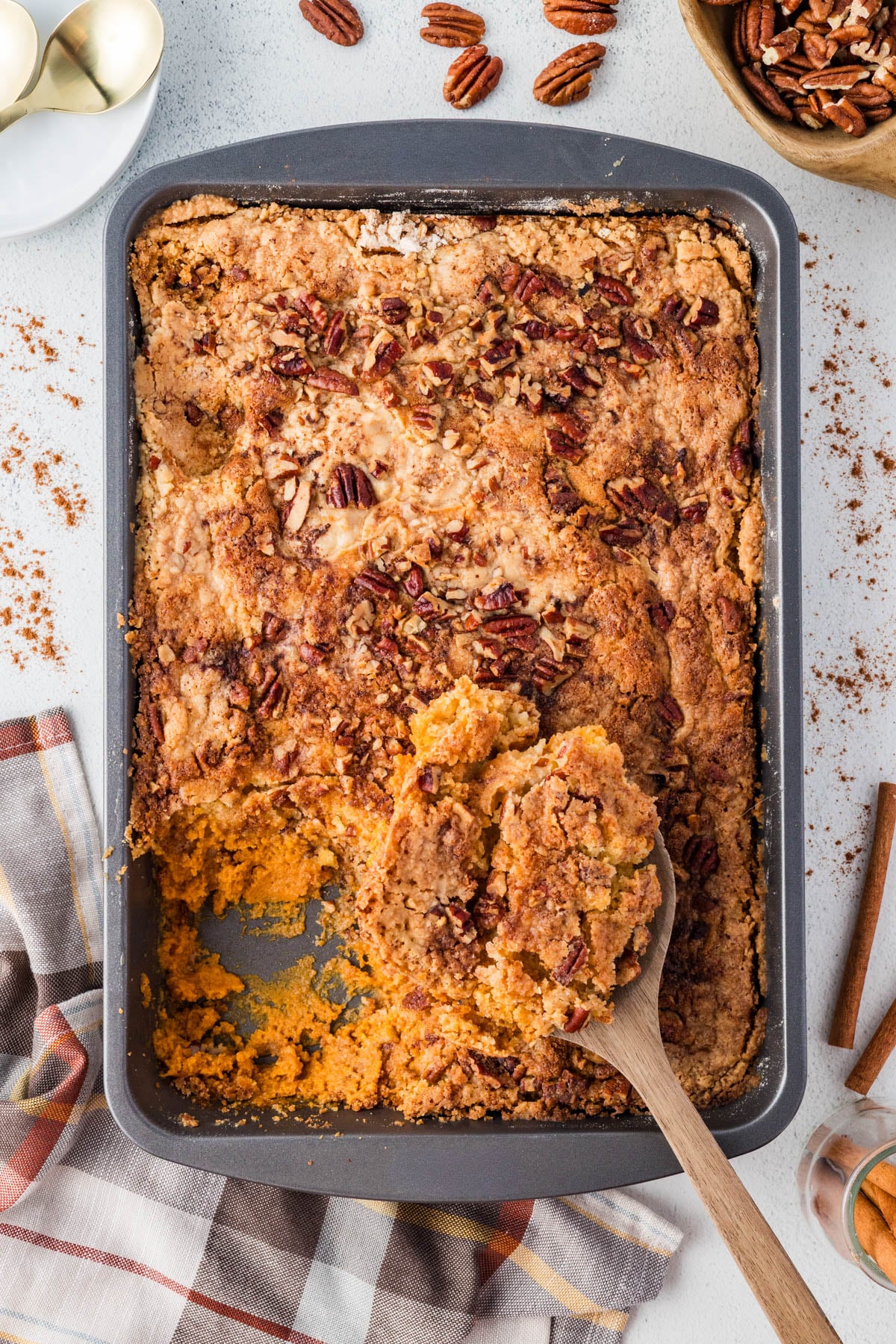 A baking pan filled with a baked dessert topped with pecans, with a portion scooped out and a wooden spoon resting in the missing section.