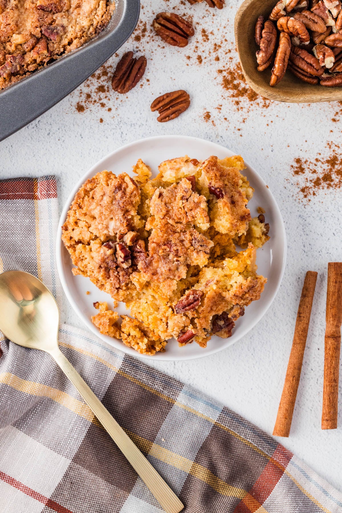 A plate of pecan crumble cake sits on a white surface near a gold spoon, a plaid napkin, cinnamon sticks, and a bowl of pecans.