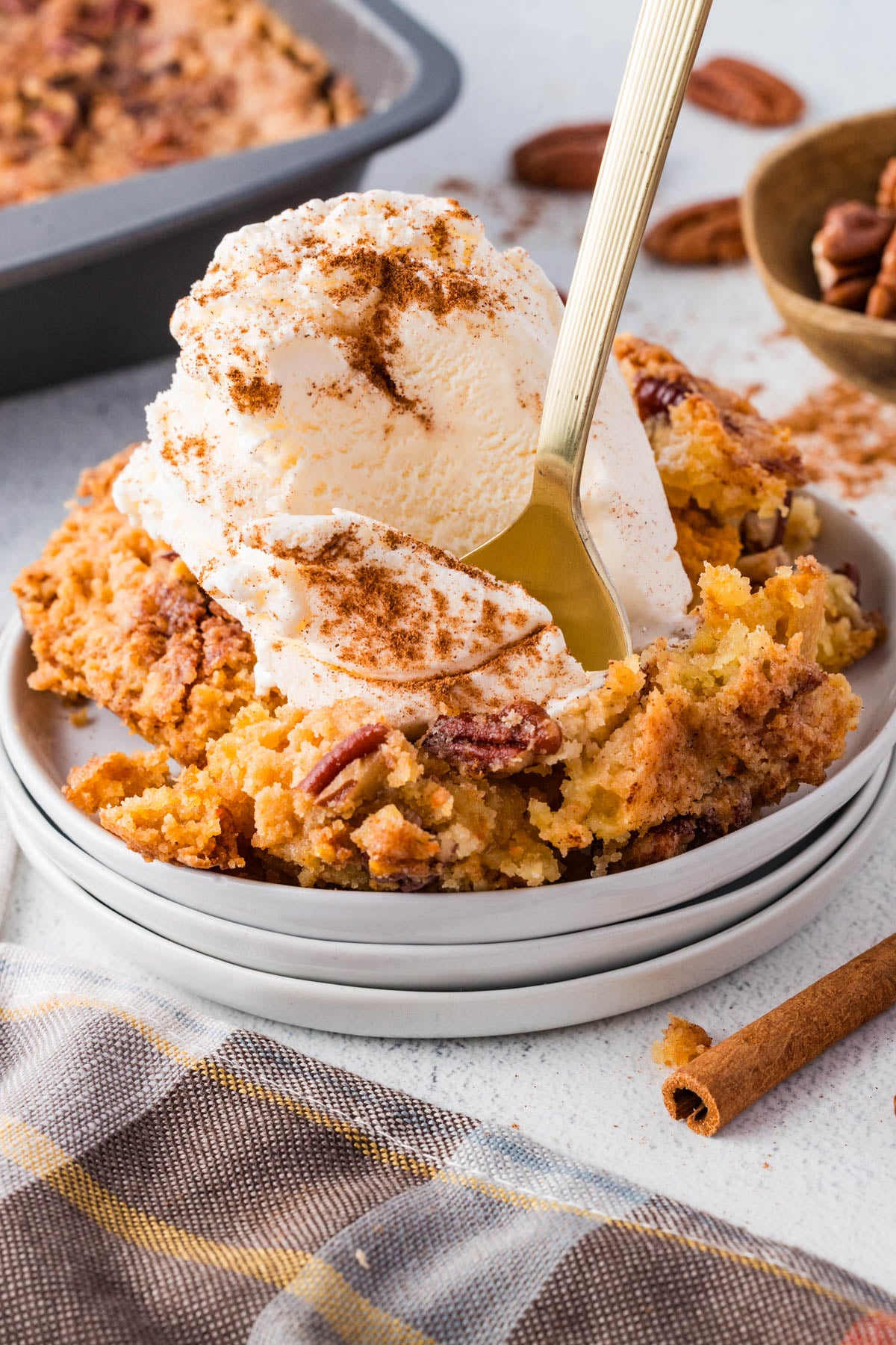 A serving of pecan dessert topped with a scoop of vanilla ice cream and cinnamon, with a fork on a white plate next to a plaid napkin.