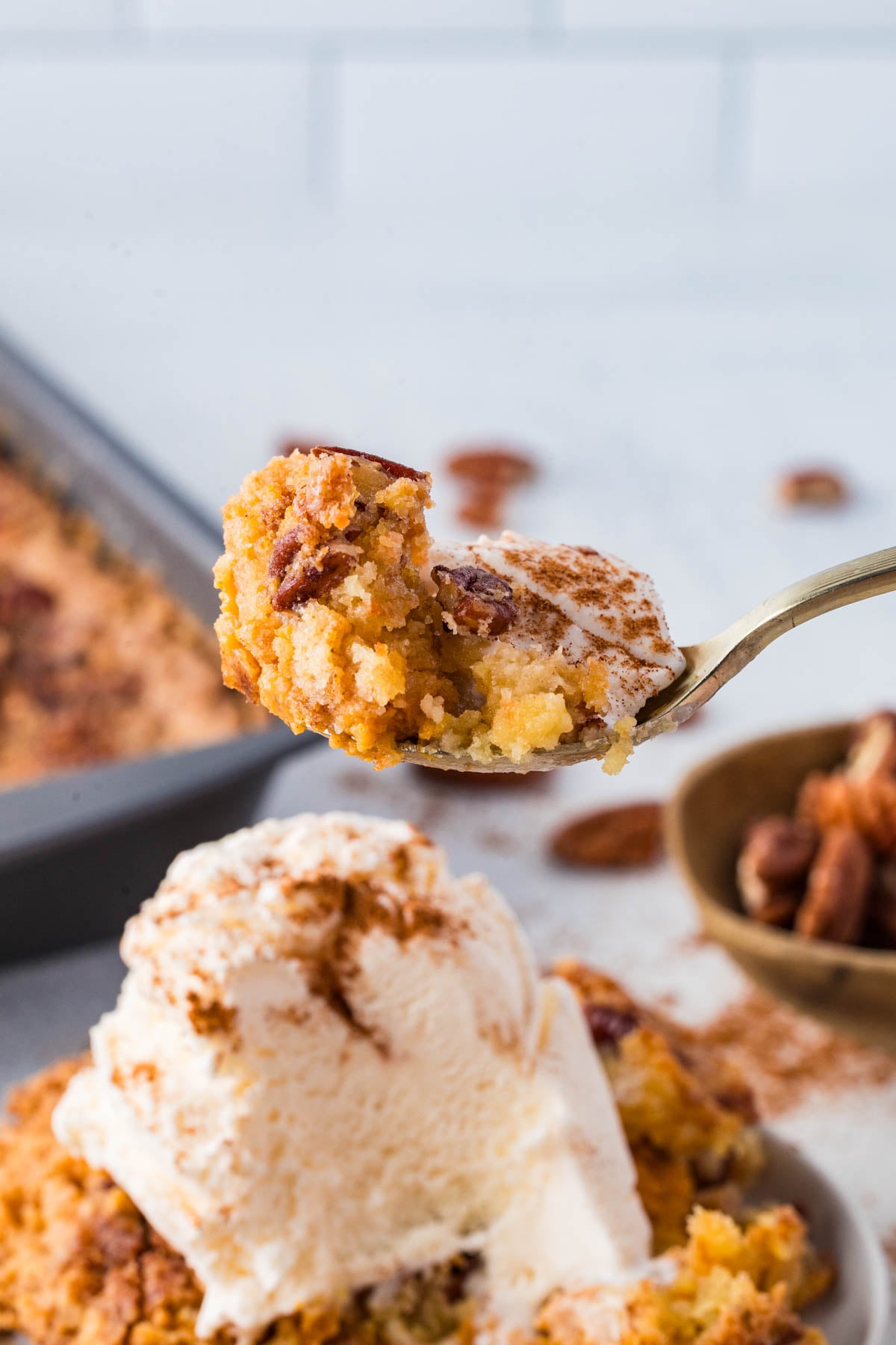 A spoonful of pumpkin dessert with pecans held above a plate, topped with a scoop of vanilla ice cream and a sprinkle of cinnamon.
