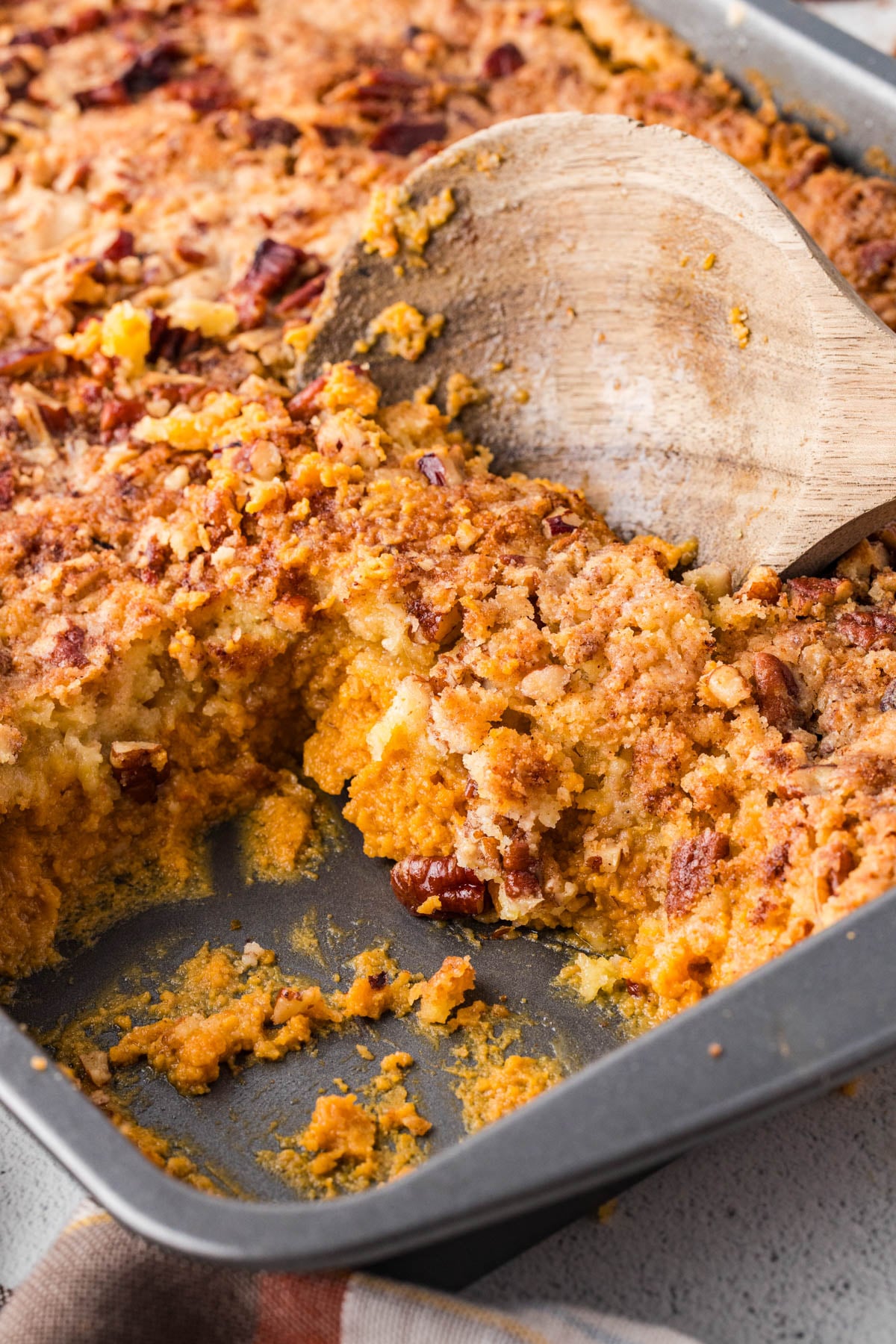 A baking pan filled with a crumbly, golden-brown pumpkin dump cake topped with chopped pecans, with a wooden spatula serving a portion.
