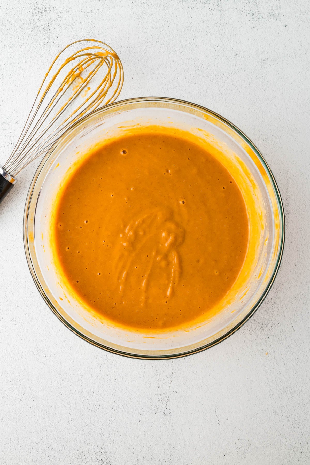 A glass bowl filled with orange batter sits on a light surface next to a metal whisk with some batter on it.