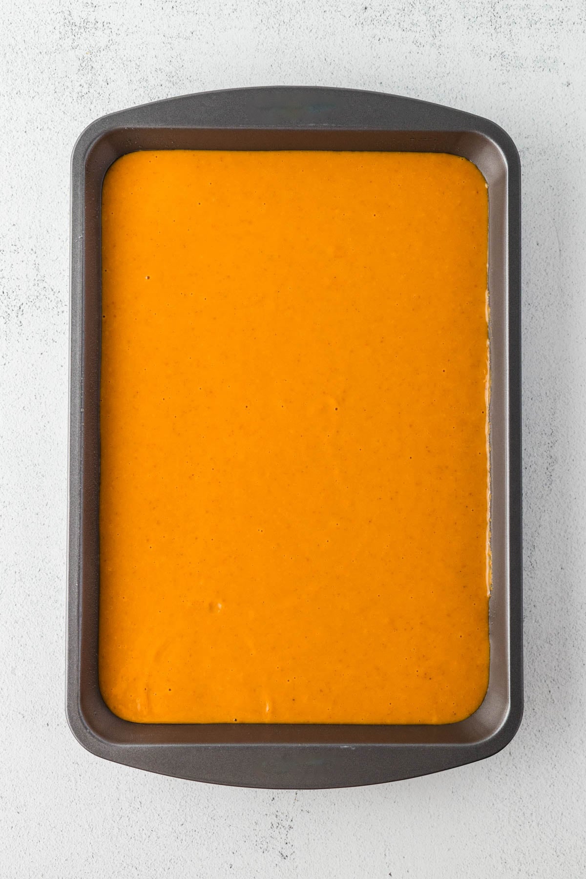A rectangular baking pan filled with an even layer of pumpkin dump cake batter, viewed from above on a light gray surface.