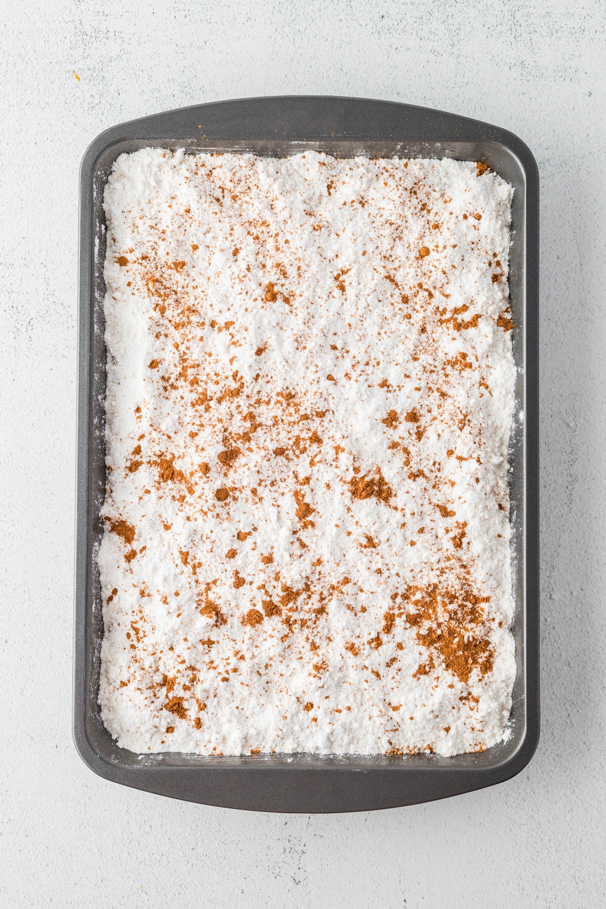 A rectangular baking pan filled with a pumpkin mixture with cake mix and a sprinkle of pumpkin pie spice on a light surface.
