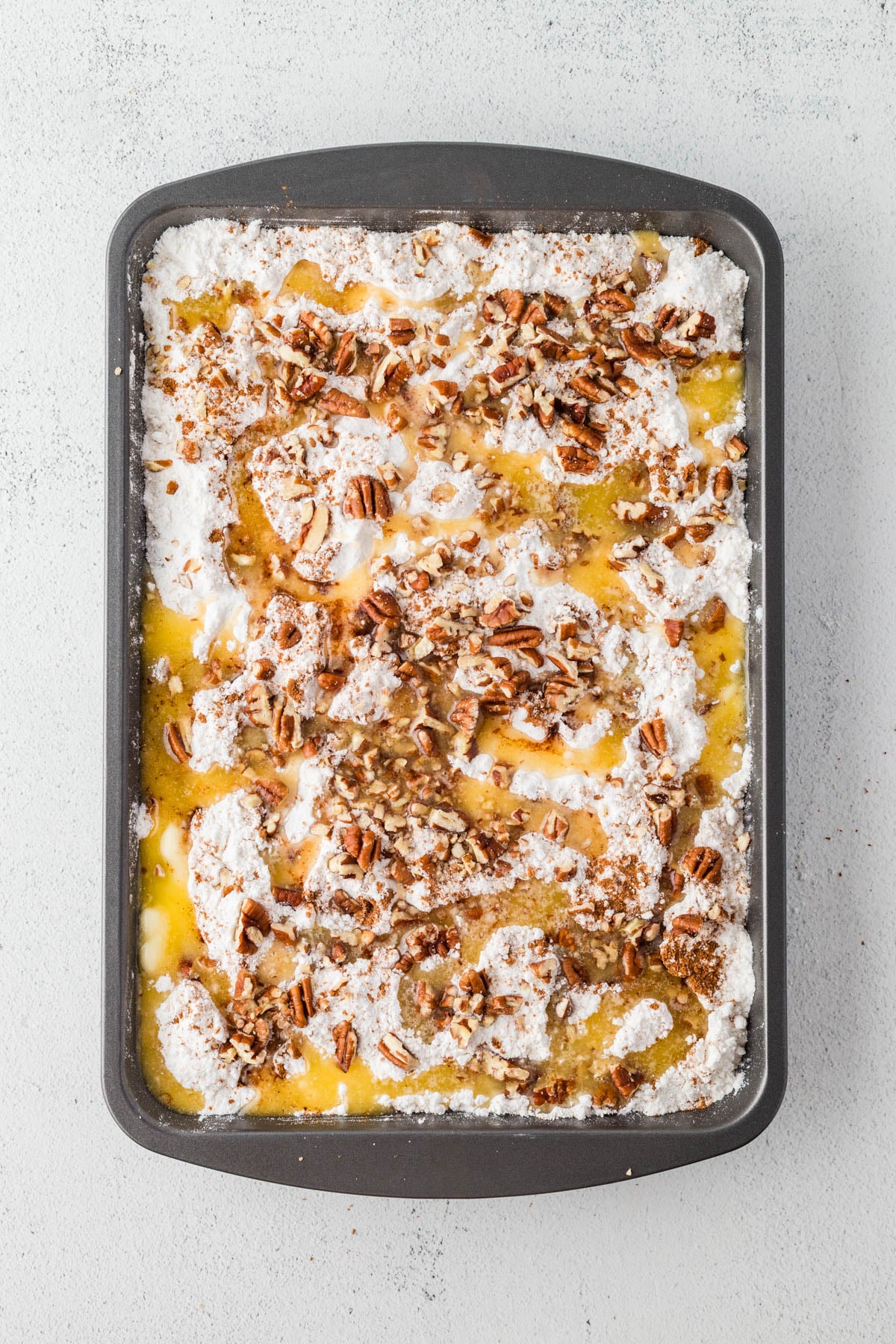 Rectangular baking pan filled with an unbaked dessert made of pumpkin batter, cake mix, chopped pecans, and melted butter, on a light surface.
