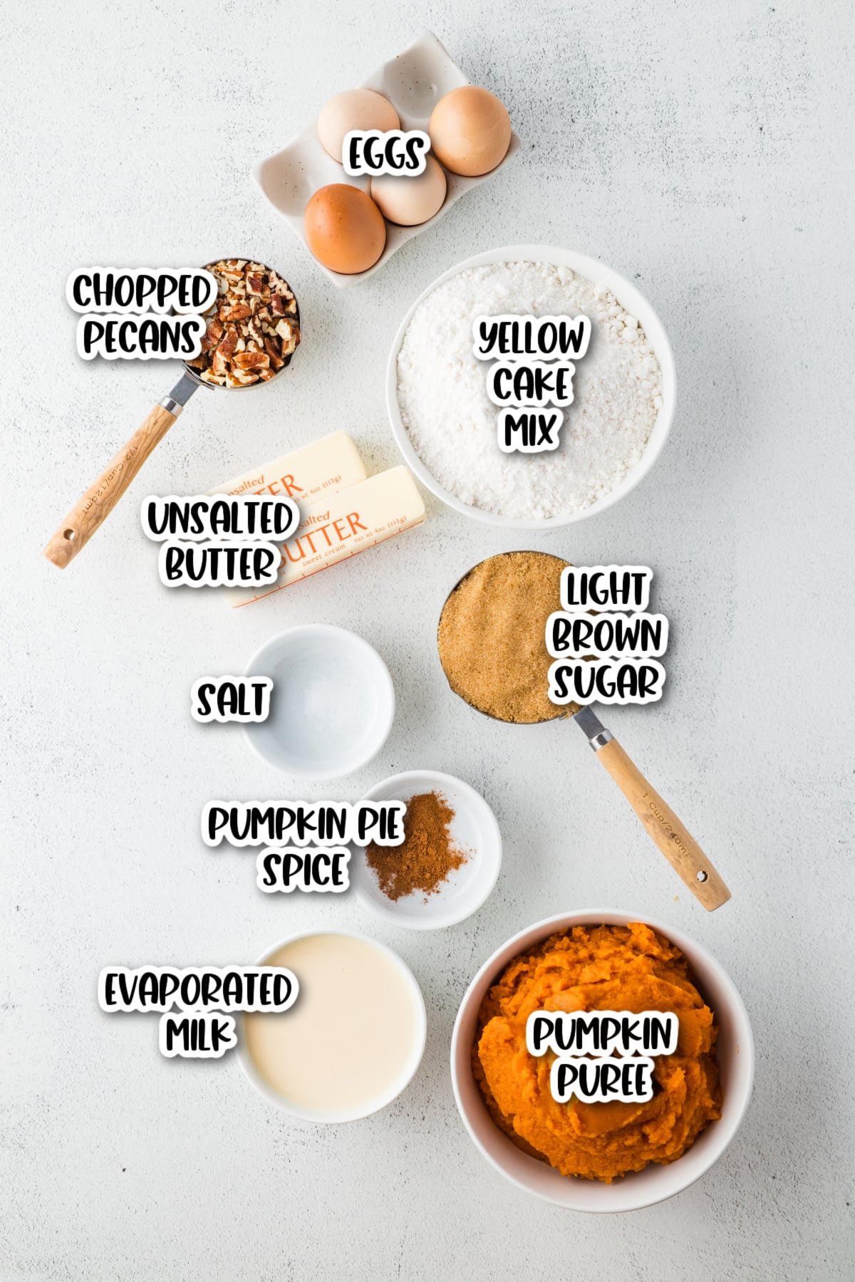 Baking ingredients arranged on a white surface, including eggs, yellow cake mix, chopped pecans, unsalted butter, light brown sugar, salt, pumpkin pie spice, evaporated milk, and pumpkin purée.