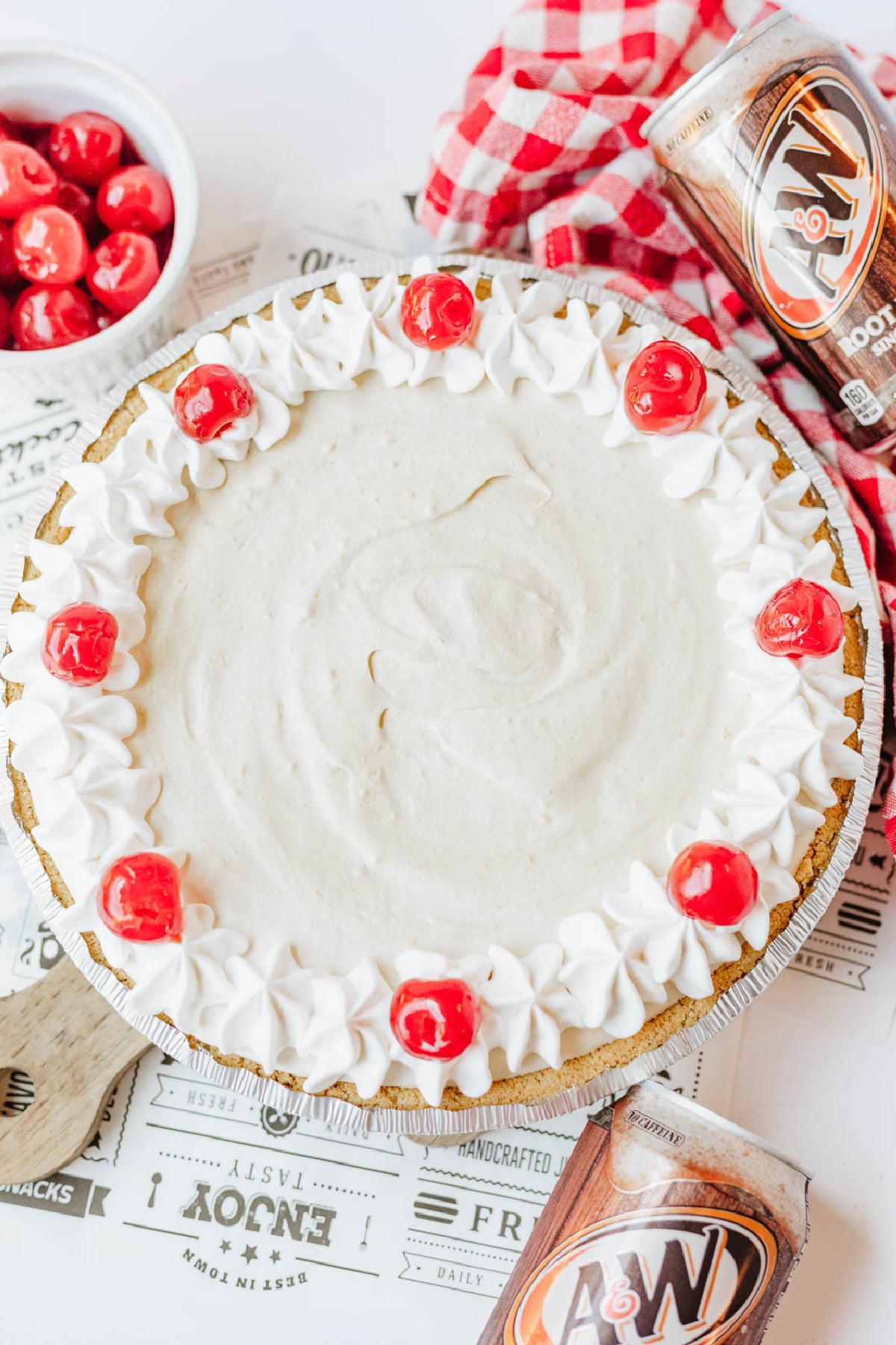 A cream pie in a graham cracker crust is topped with whipped cream swirls and cherries, with cans of A&W root beer and a bowl of cherries nearby.