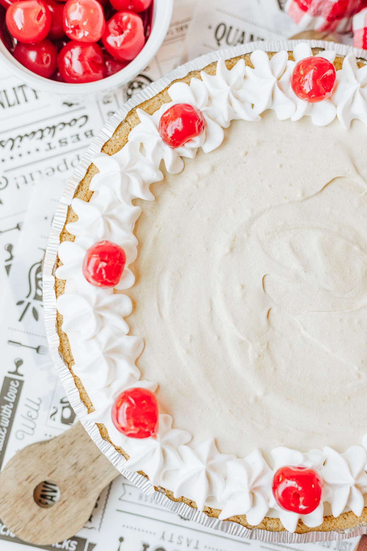 Root beer float pie topped with Cool Whip swirls and maraschino cherries, next to a bowl of cherries on a printed paper background.