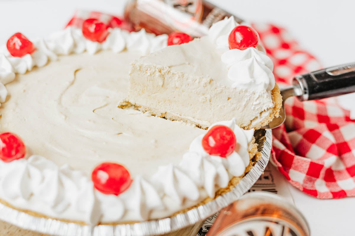 A root beer float pie with a graham crust topped with Cool Whip and cherries, with one slice being lifted out on a pie server.