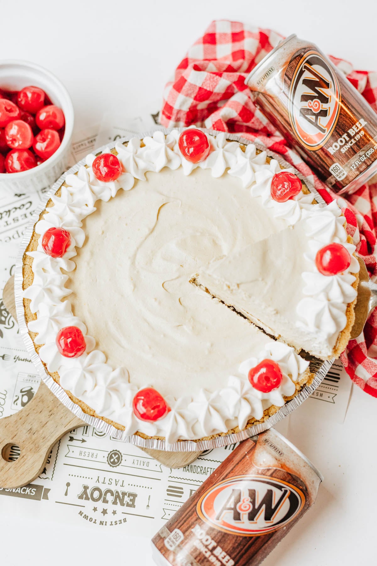 A root beer float pie topped with Cool Whip and cherries, with one slice missing. A can of A&W root beer and a bowl of cherries are nearby on a red checkered cloth.