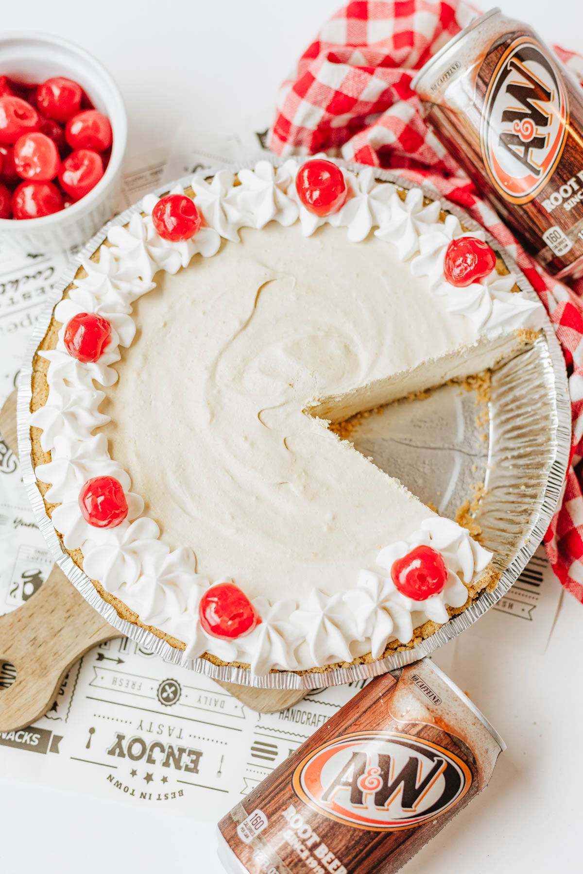 A cream pie topped with Cool Whip and cherries, with one slice missing, sits next to A&W root beer cans and a bowl of cherries on a white surface.