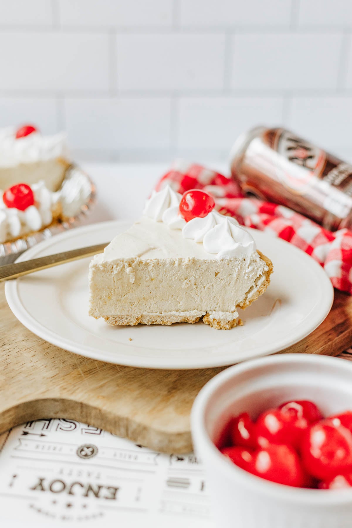A slice of cream pie with Cool Whip and a cherry on a white plate, with more pie and cherries in the background.