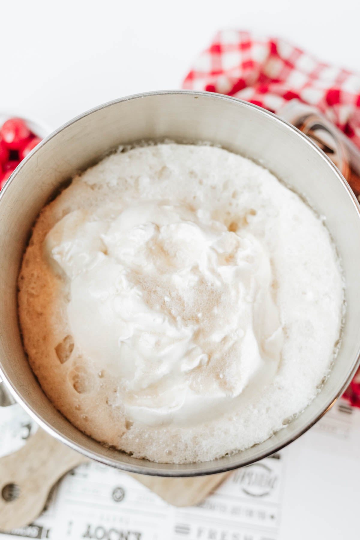 A mixing bowl filled with a mixture of root beer and Cool Whip sits on a countertop with a red checkered cloth and kitchen utensils nearby.