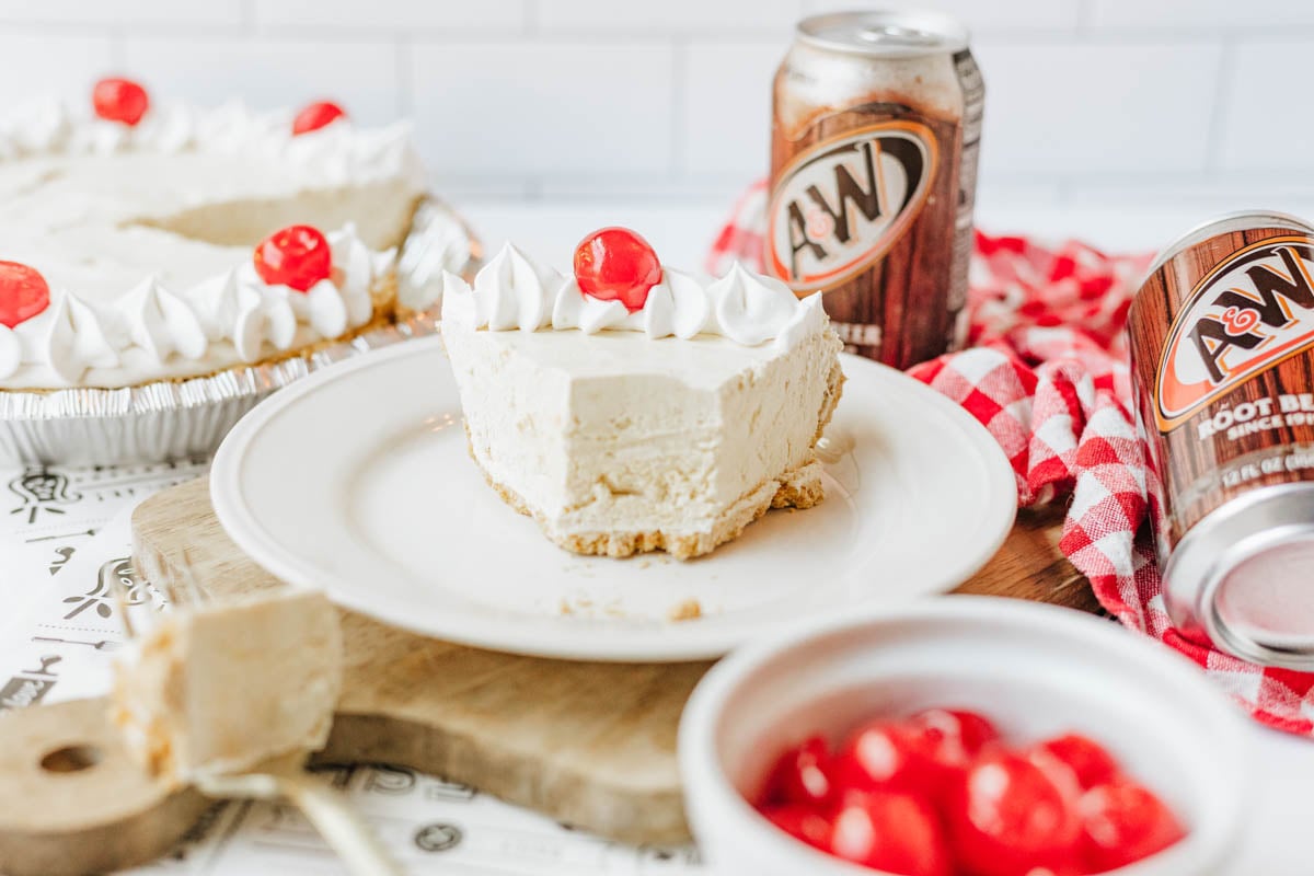 A slice of root beer float pie with a cherry on top and a bite taken out sits on a plate, surrounded by cans of A&W root beer and a bowl of cherries. The rest of the pie is in the background.
