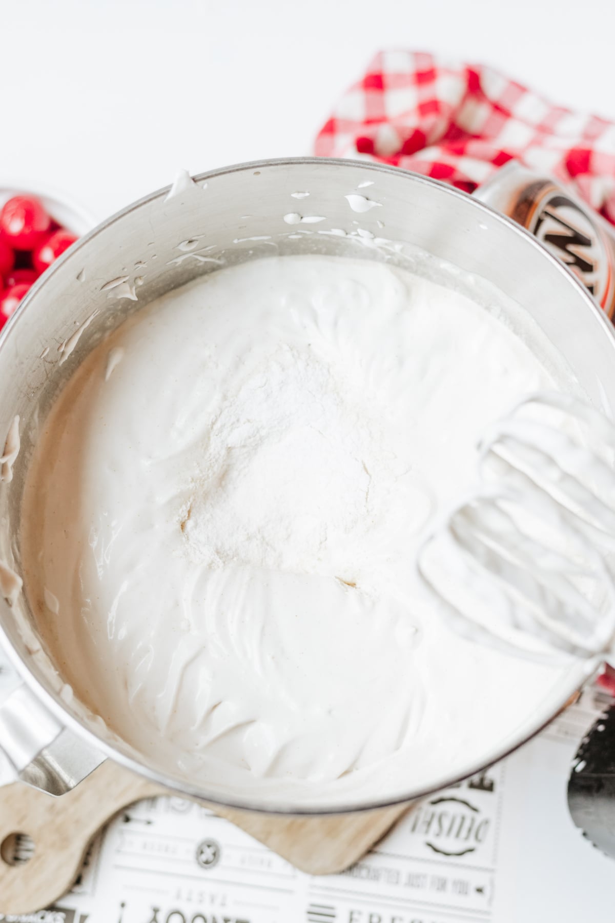 A metal mixing bowl filled with a Cool Whip mixture and vanilla pudding in the center, beside a metal hand mixer and a red checkered cloth.