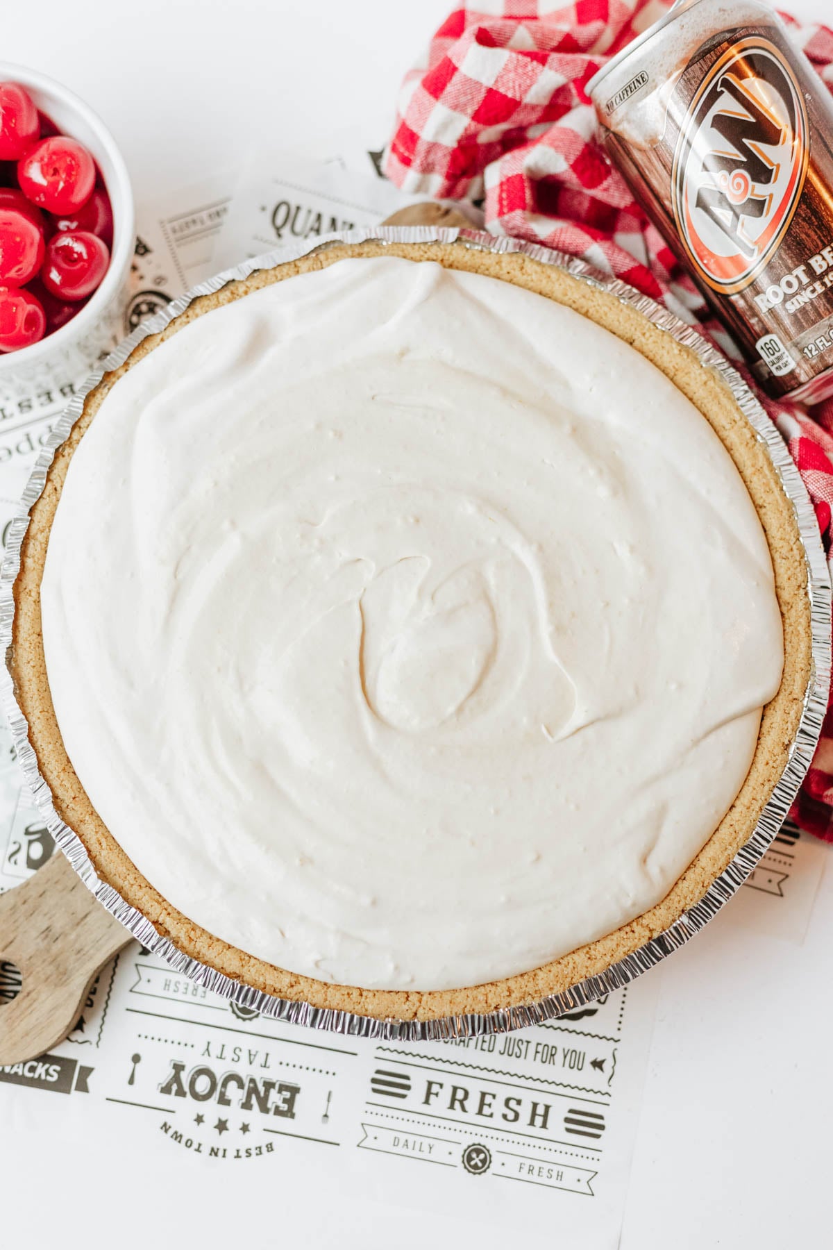 A no-bake cream pie in a graham cracker crust sits on newspaper print, with a can of root beer, red checkered cloth, and a bowl of maraschino cherries nearby.