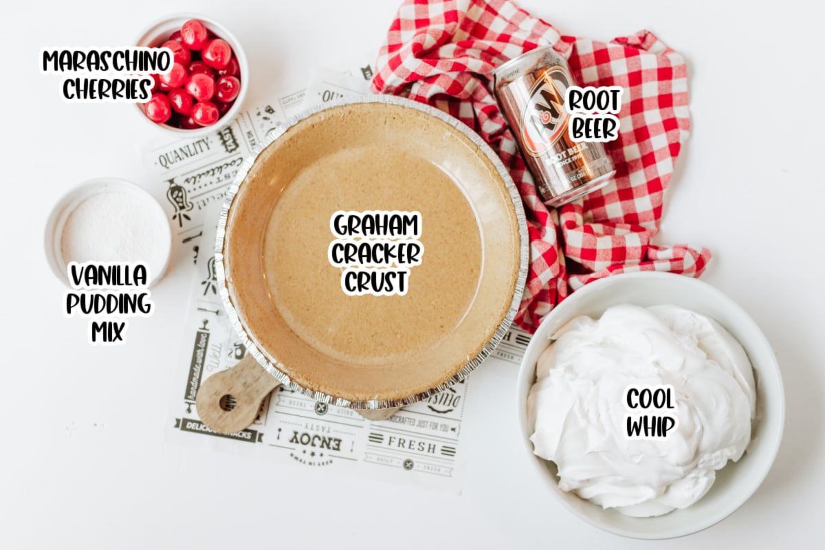 A graham cracker crust, bowl of Cool Whip, vanilla pudding mix, maraschino cherries, and can of root beer are arranged on a table with a red checkered cloth.