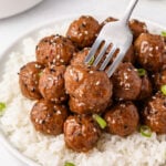A fork lifts a glazed meatball from a plate of white rice topped with several meatballs, black and white sesame seeds, and sliced green onions.