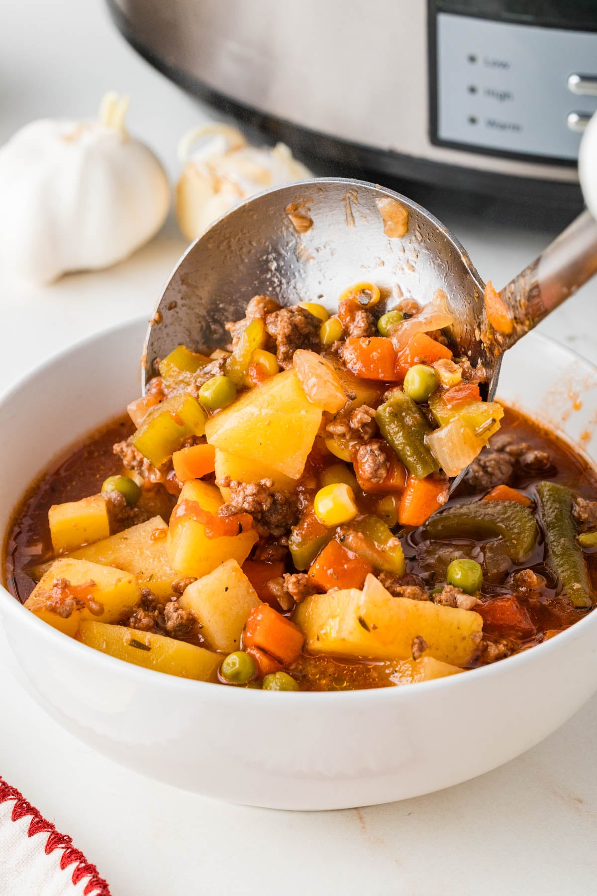 A ladle serves a bowl of hamburger soup with potatoes, carrots, green beans, and peas, in front of a slow cooker and garlic bulbs.