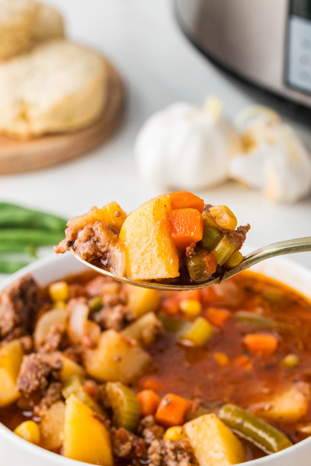 A spoonful of hamburger soup with potatoes, carrots, celery, and corn held above a bowl, with biscuits and garlic in the background.