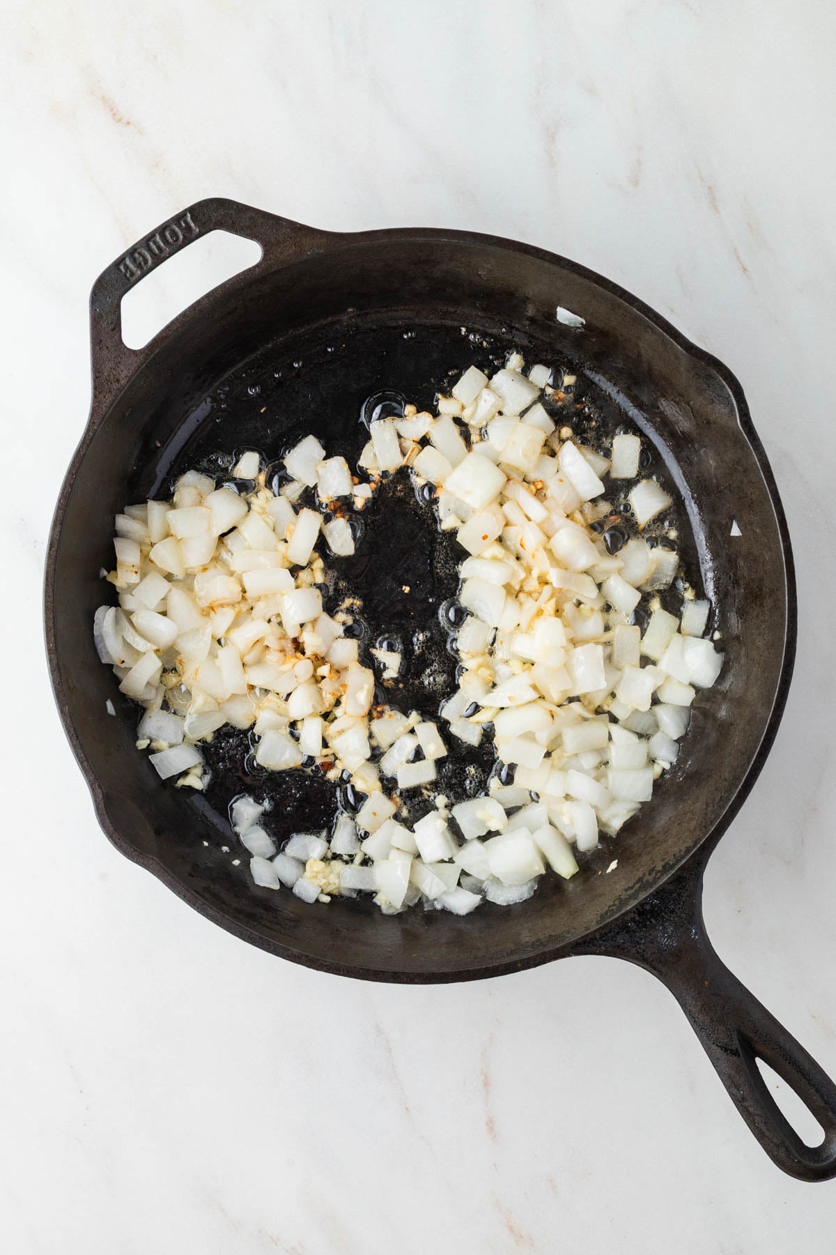 Chopped onions and minced garlic sautéing in oil in a cast iron skillet on a white marble surface.