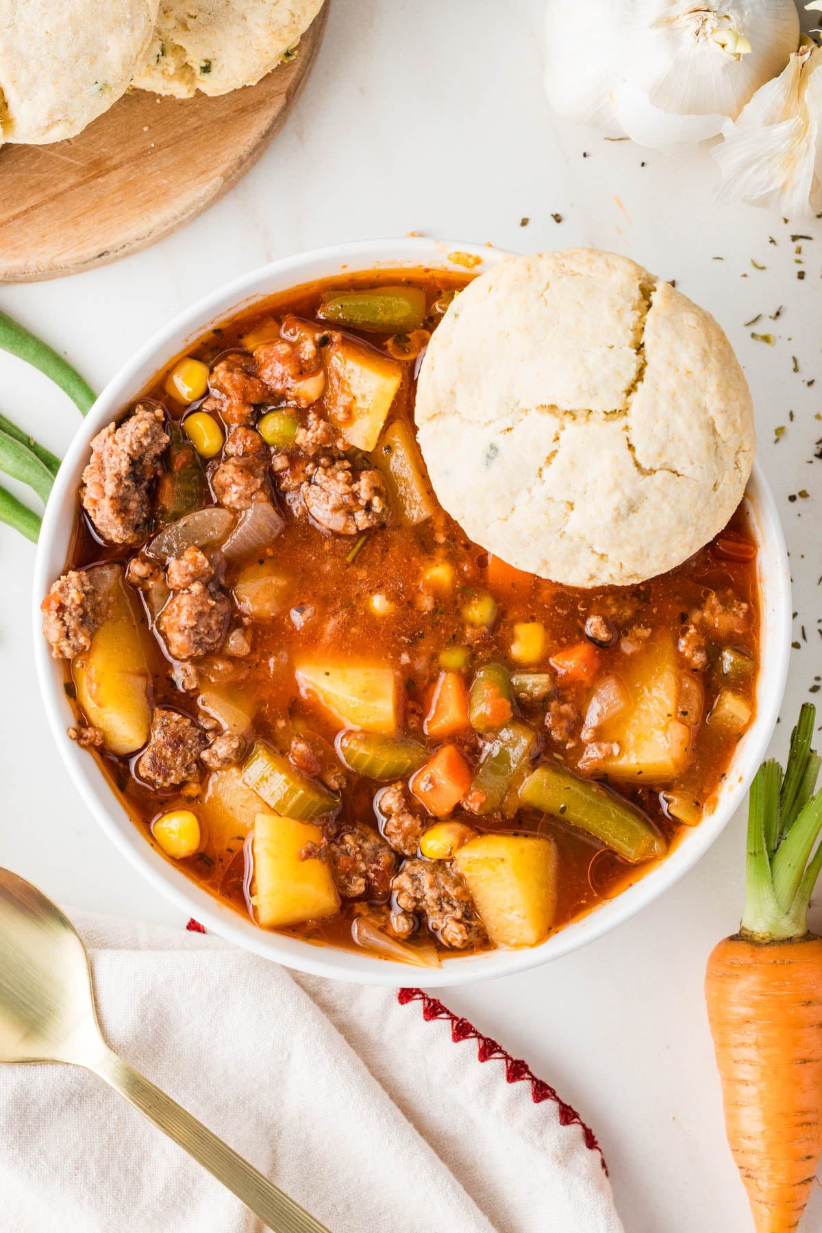 A bowl of slow cooker hamburger soup with potatoes, carrots, corn, and peas, topped with a biscuit, next to a spoon, carrot, green onions, and garlic.