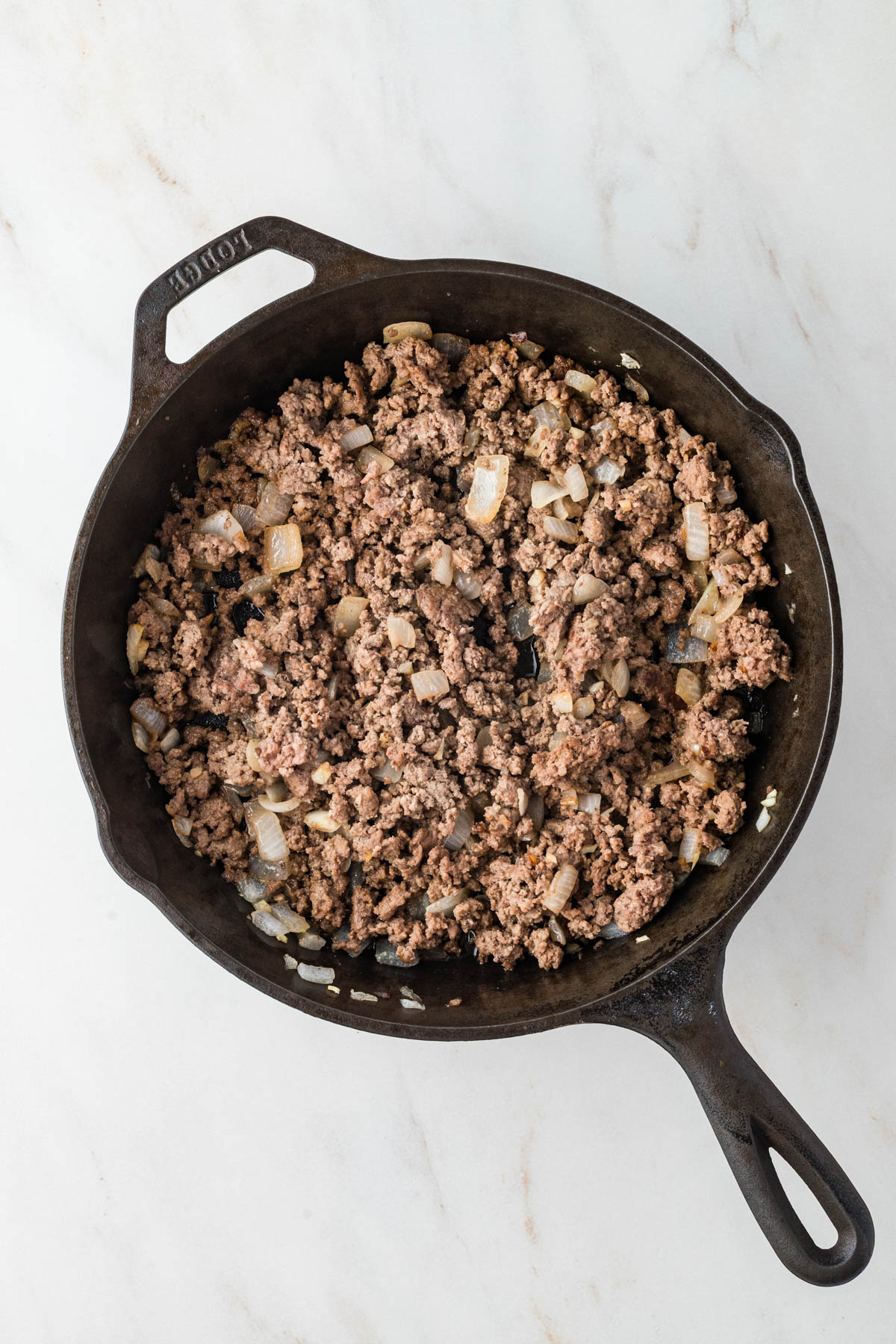 Cooked ground beef and diced onions in a black cast iron skillet on a white marble surface.
